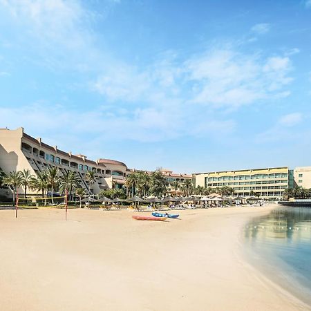 Al Raha Beach Resort & Spa Abu Dhabi Exterior foto The photo depicts a serene beach scene with a clear blue sky. In the foreground, there is a sandy beach that gently slopes down to calm water. On the left side, several palm trees provide a tropical atmosphere. In the background, there are two large 