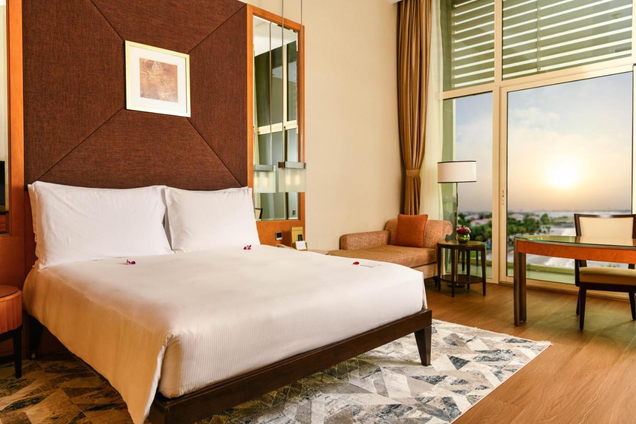 Al Raha Beach Resort & Spa Abu Dhabi Exterior foto The photo shows a stylish hotel room featuring a large bed with white bedding, positioned against a textured wall. There’s a small seating area with a chair and a table, and a window with curtains that allows natural light to fill the space. The room