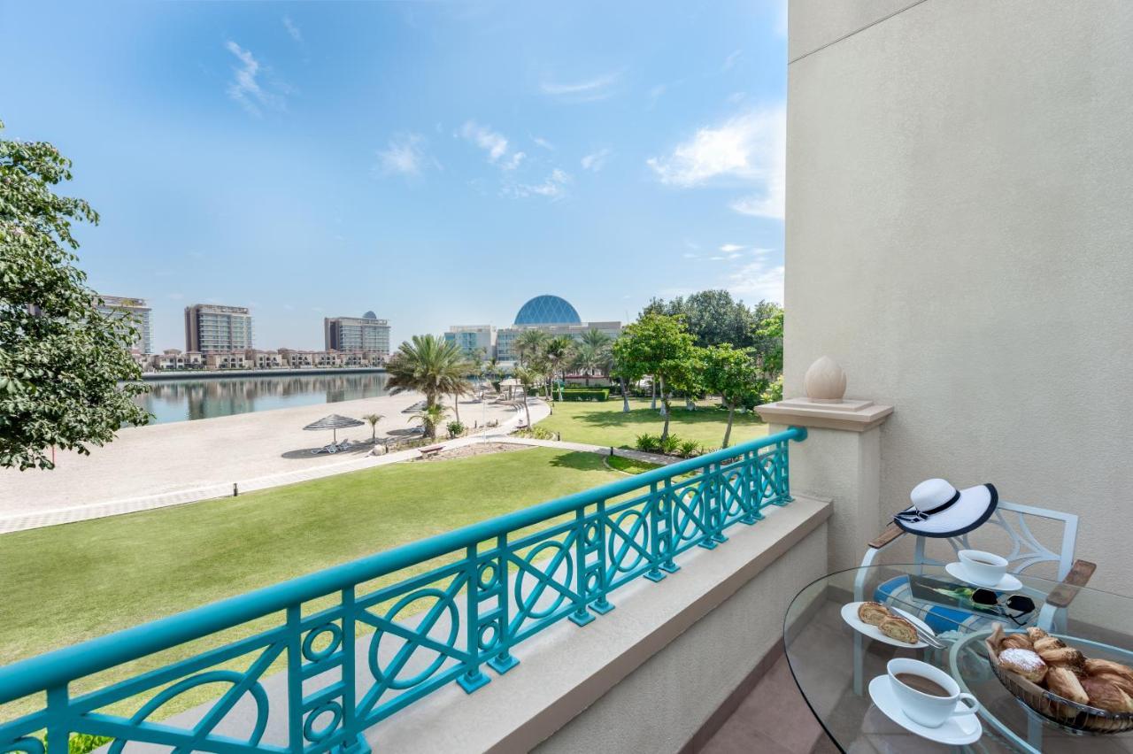 Al Raha Beach Resort & Spa Abu Dhabi Exterior foto The photo shows a balcony view overlooking a scenic landscape. In the foreground, there is a glass table set with snacks or pastries and two cups. Beyond the balcony railing, there is a well-maintained green area with palm trees and a pathway. The ba