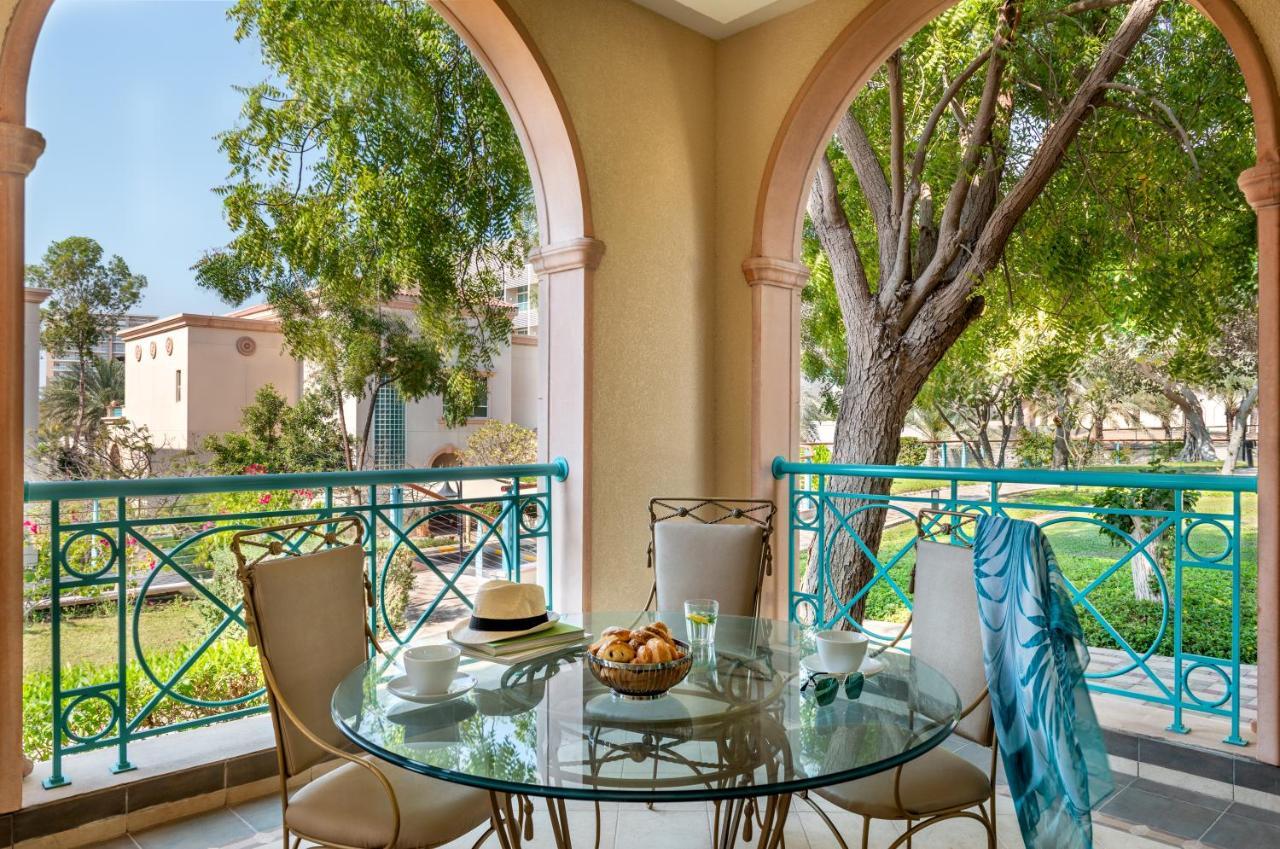 Al Raha Beach Resort & Spa Abu Dhabi Exterior foto The photo shows a serene outdoor balcony setting. There is a circular glass table with a bowl of fruit or snacks in the center, alongside a glass and a small plate. Surrounding the table are four elegant chairs. The railing is turquoise, framing a vi