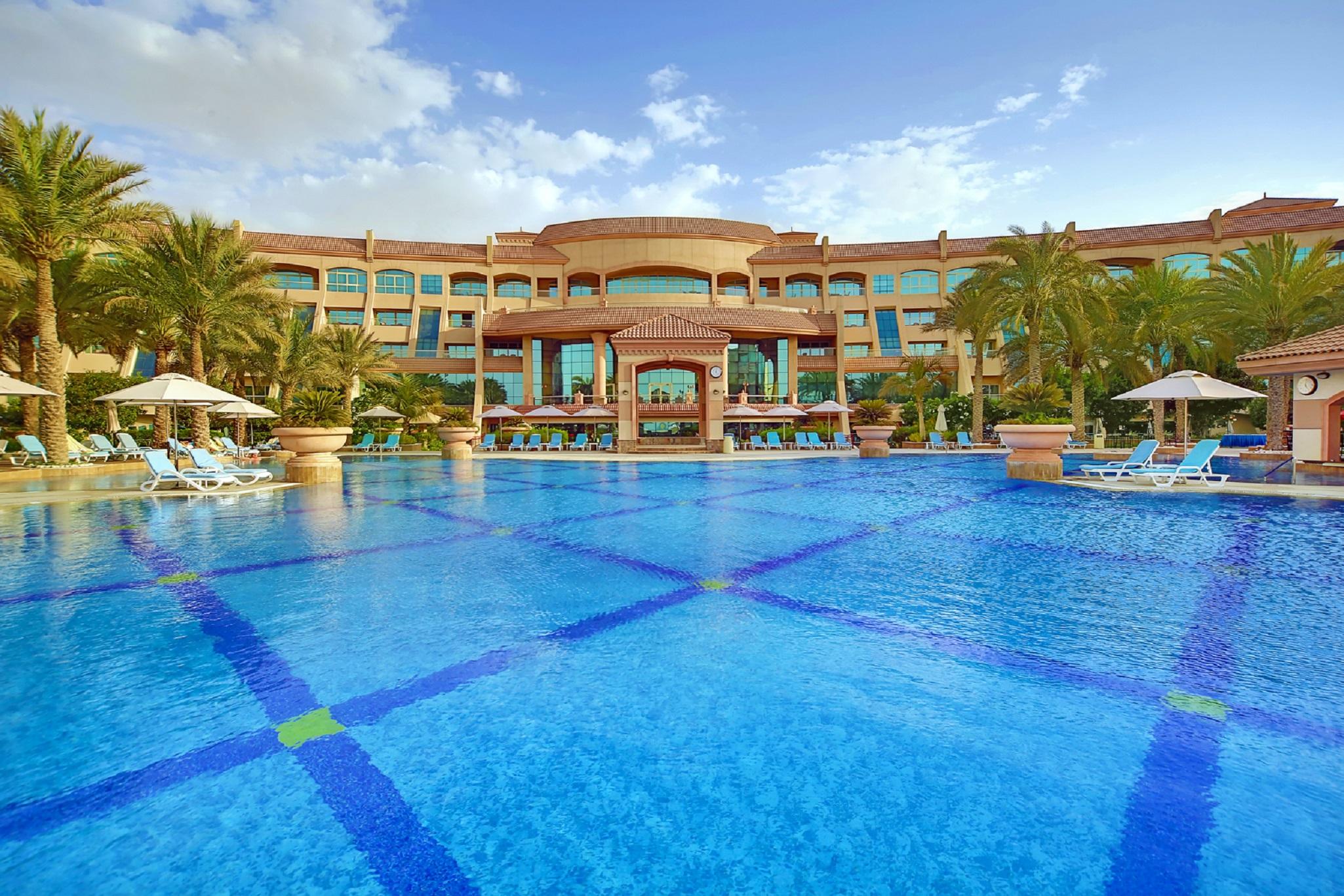 Al Raha Beach Resort & Spa Abu Dhabi Facilități foto The photo features a luxurious resort with a large swimming pool in the foreground. The pool is crystal clear and reflects the sky, bordered by lounge chairs and palm trees. In the background, the resort building showcases modern architecture with la