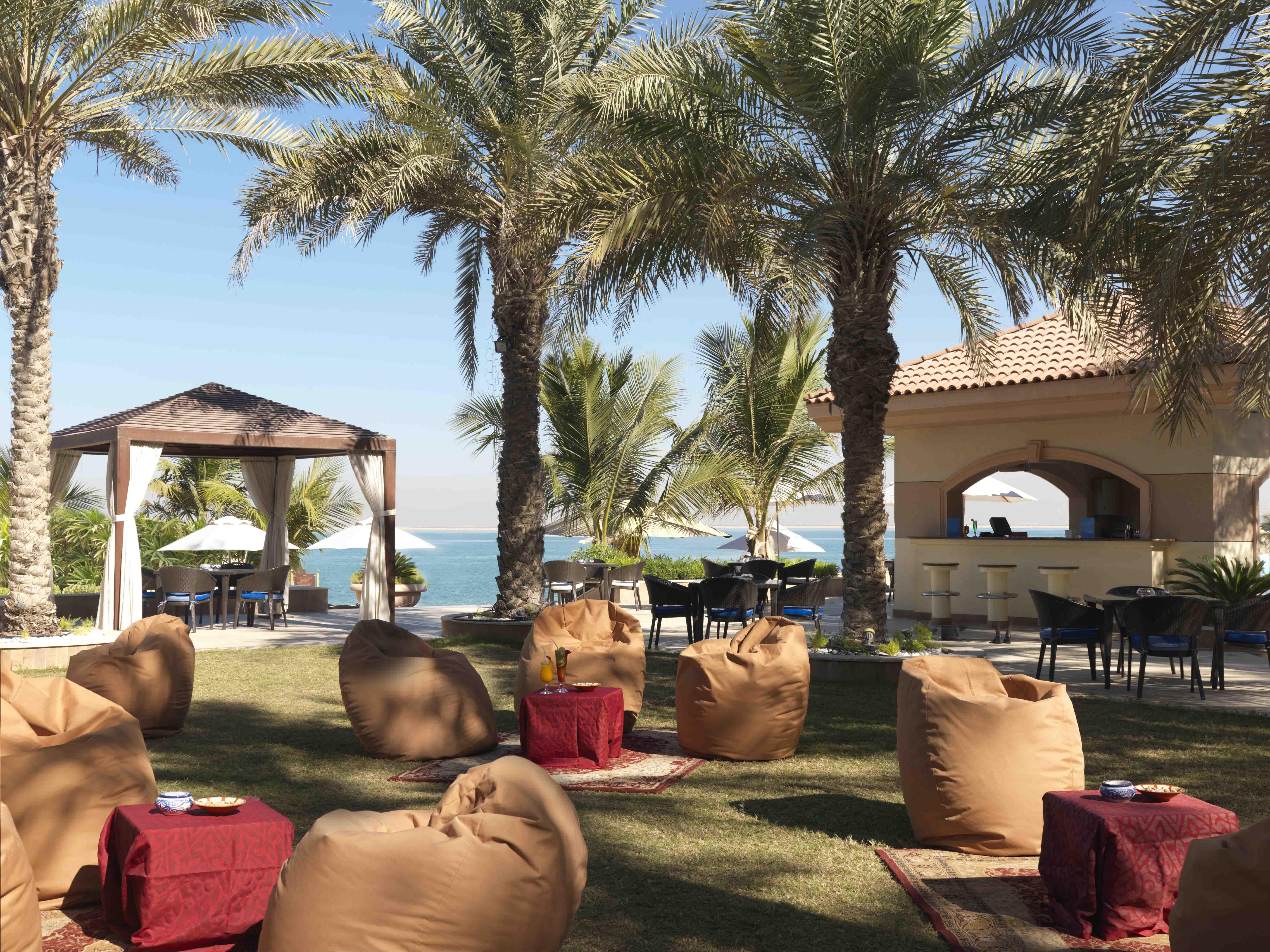 Al Raha Beach Resort & Spa Abu Dhabi Restaurant foto The photo depicts a beautiful outdoor setting with several elements of relaxation and leisure. In the foreground, there are large, comfortable bean bag chairs arranged on a grassy area, accompanied by low tables. Surrounding this seating area are tal