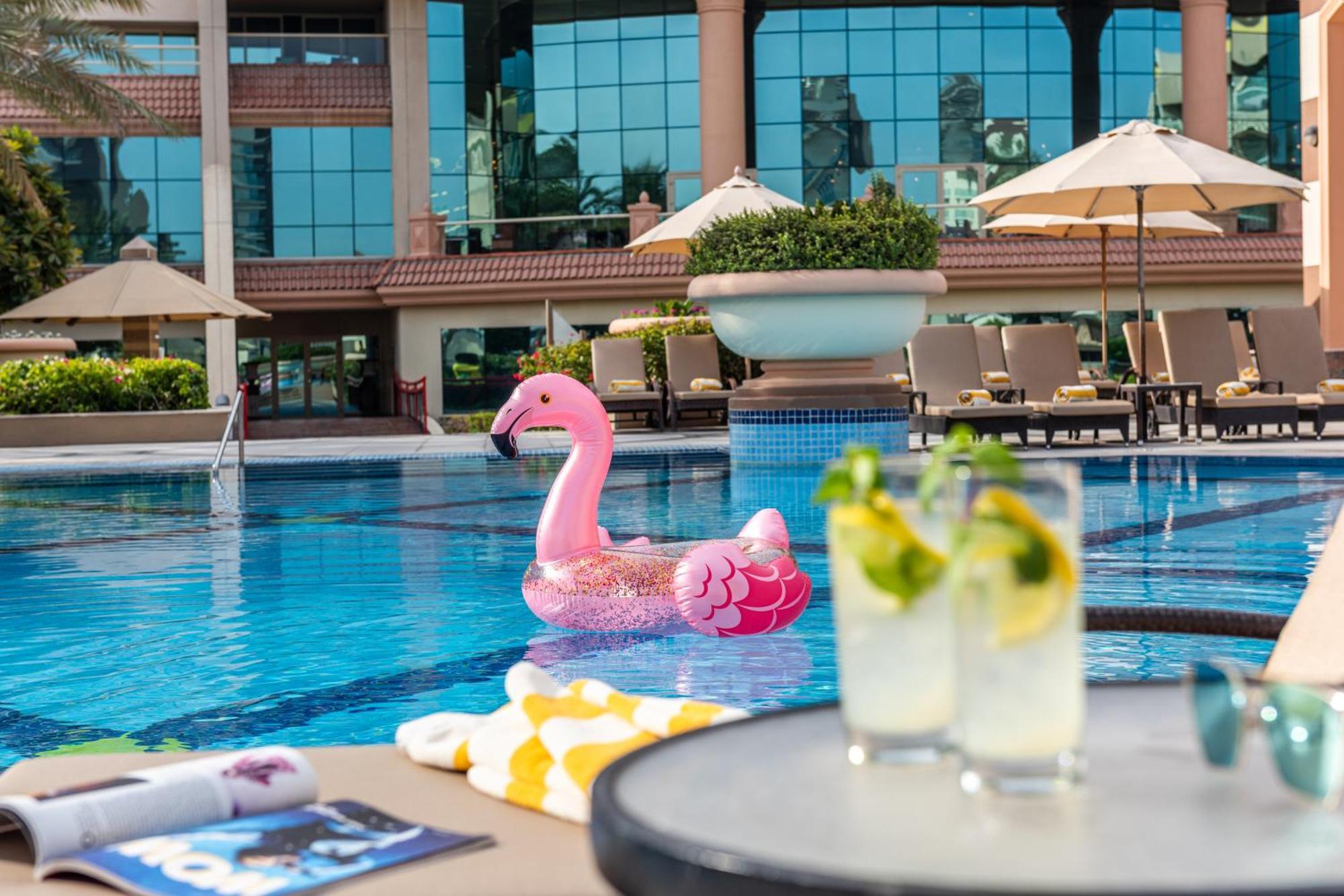 Al Raha Beach Resort & Spa Abu Dhabi Exterior foto The photo depicts a bright and inviting outdoor swimming pool surrounded by a luxurious setting. In the pool, there is a large pink flamingo float, adding a fun and whimsical touch. To the side, a table is set up with two refreshing drinks, likely co