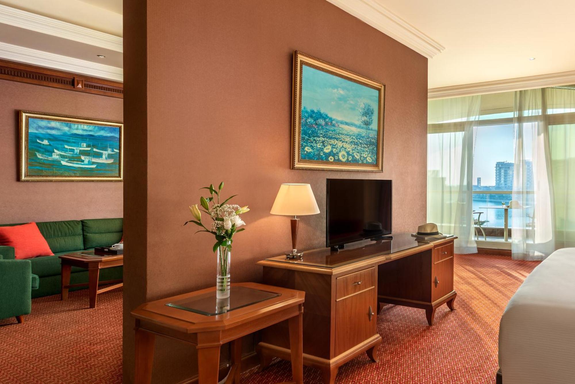 Al Raha Beach Resort & Spa Abu Dhabi Cameră foto The photo shows a hotel room with a warm, inviting atmosphere. There's a large window that provides natural light and a view, likely overlooking a cityscape or water. On one wall, there's a piece of artwork featuring a landscape, and nearby, a small 