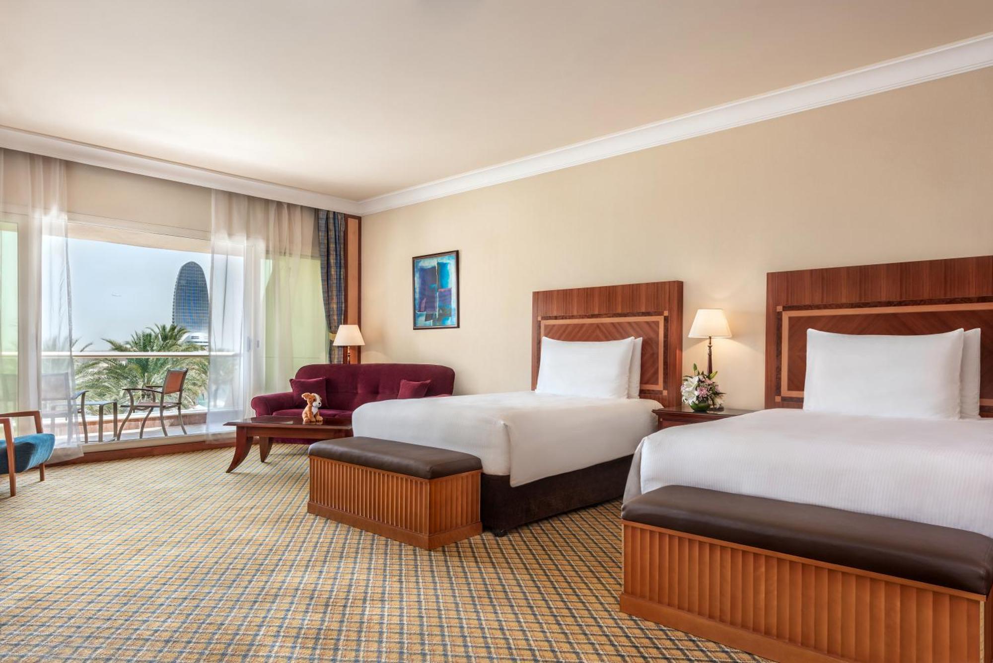 Al Raha Beach Resort & Spa Abu Dhabi Cameră foto The photo shows a hotel room featuring two twin beds with wooden headboards. The room has a light, neutral color scheme, with a large window that provides natural light and a view of the outdoors. There is a small seating area with a purple sofa and 