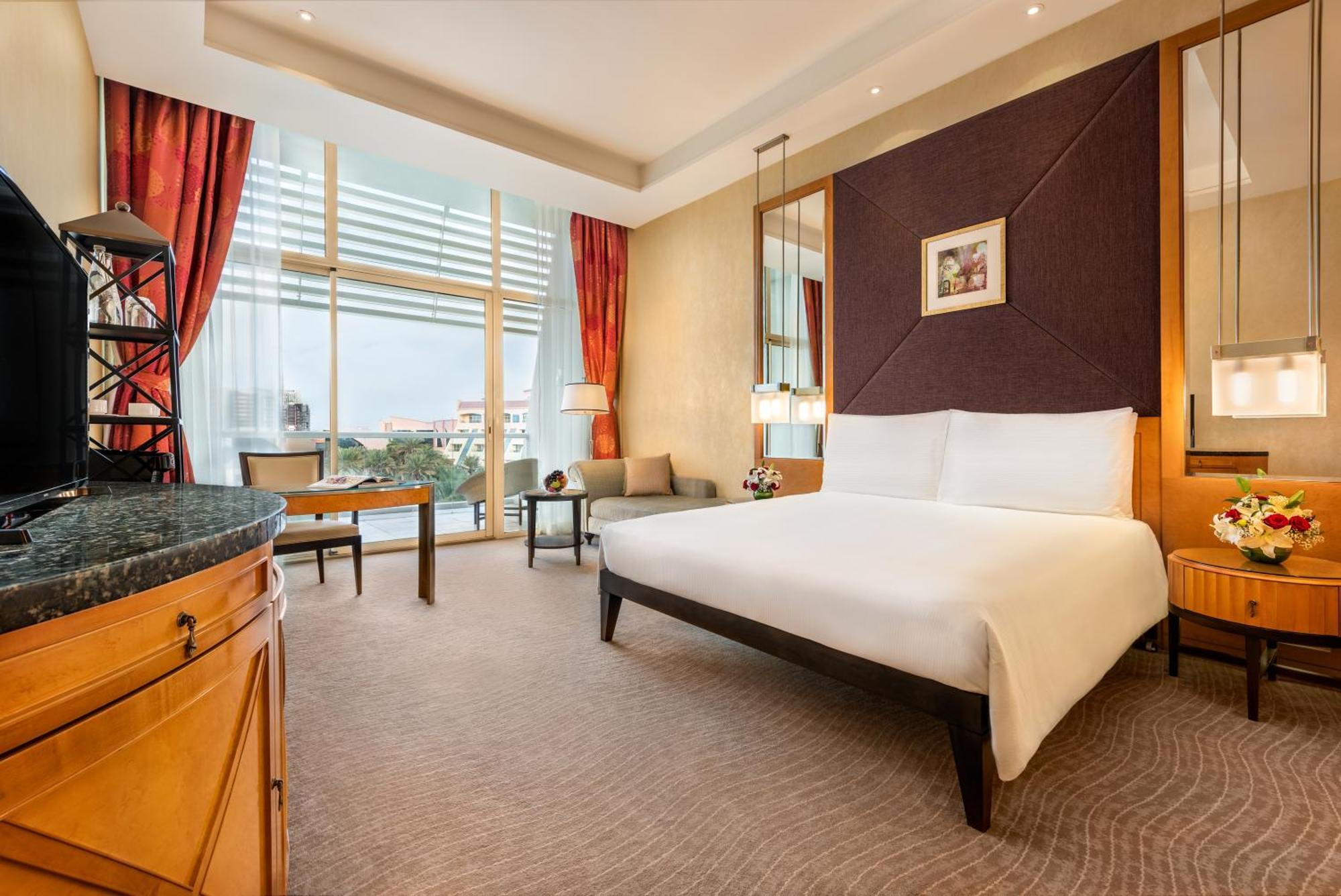 Al Raha Beach Resort & Spa Abu Dhabi Cameră foto The photo shows a modern hotel room featuring a large bed with a white duvet positioned against a purple accent wall. There are tall windows with sheer curtains allowing natural light to enter the space, along with red drapes framing the windows. A d