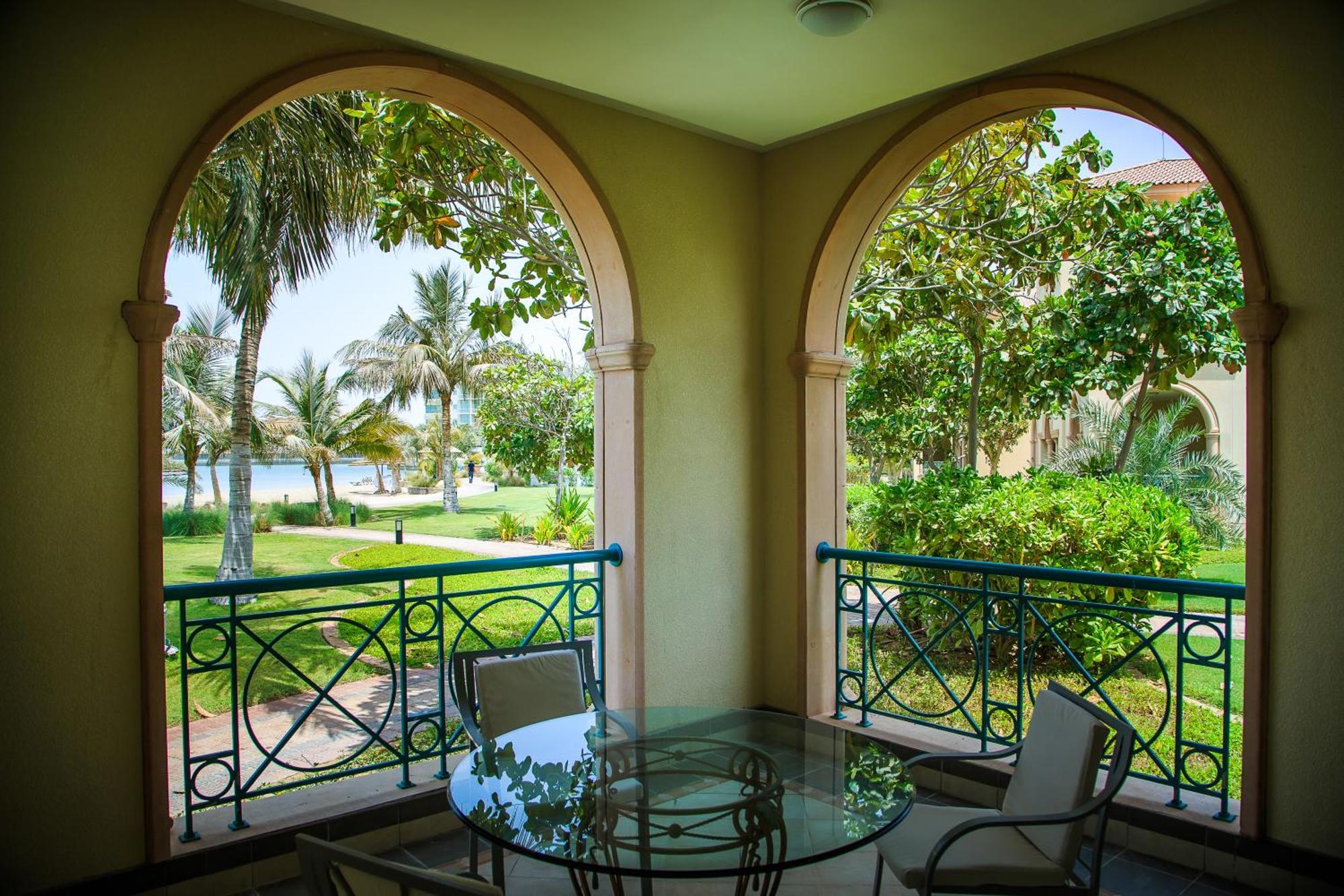 Al Raha Beach Resort & Spa Abu Dhabi Cameră foto The photo displays a beautifully designed balcony or terrace area. It features two large arches that frame the view outside. Through the arches, you can see a lush garden with palm trees and well-maintained greenery. There’s a glass table with severa